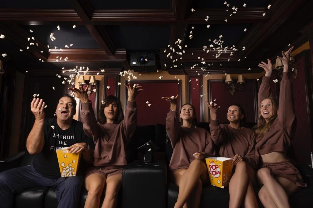 Family and friends enjoying popcorn in a dedicated home theater with black recliners, warm lighting, and a star ceiling.