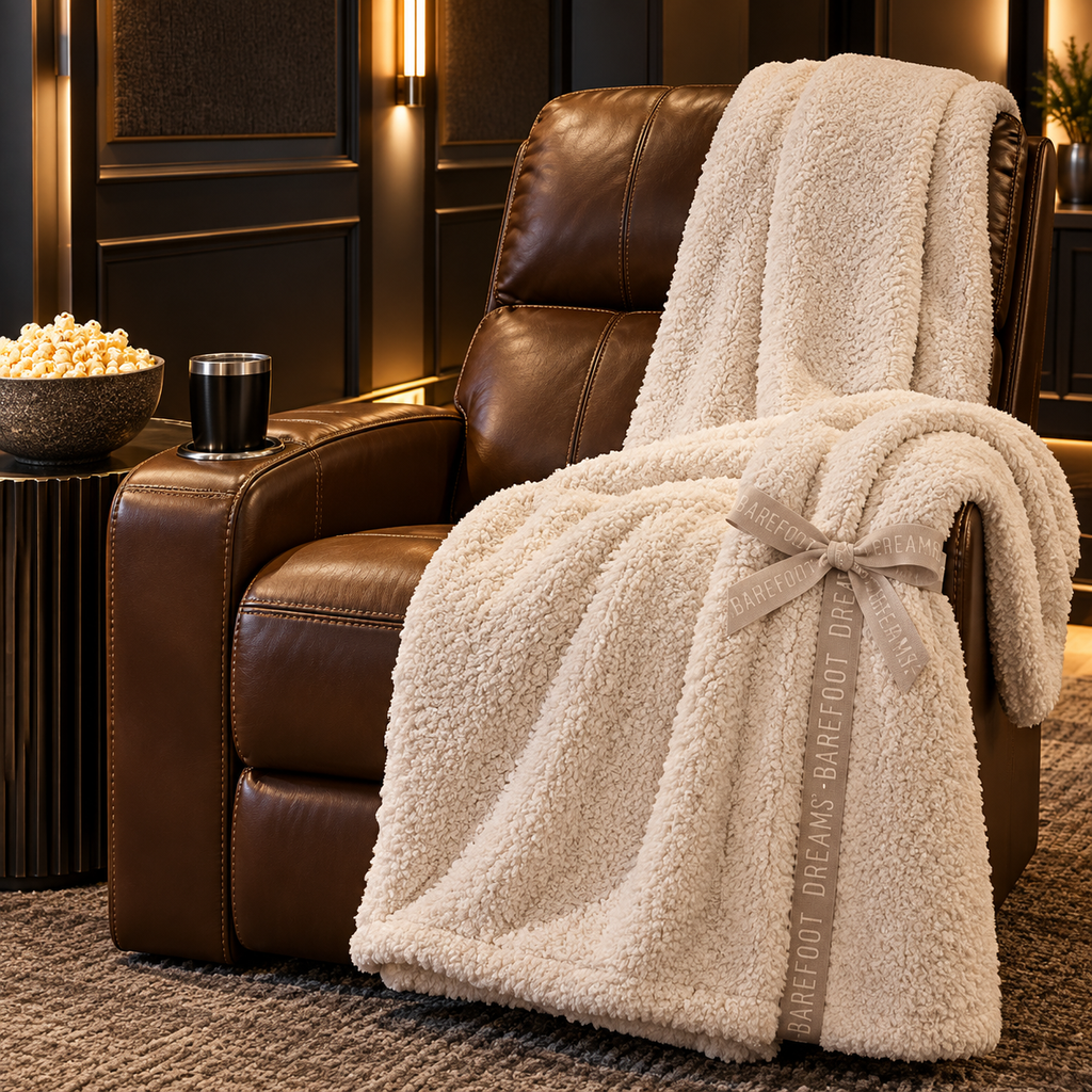 Cream Barefoot Dreams throw draped over a brown leather home theater recliner beside a bowl of popcorn in a warmly lit movie room.